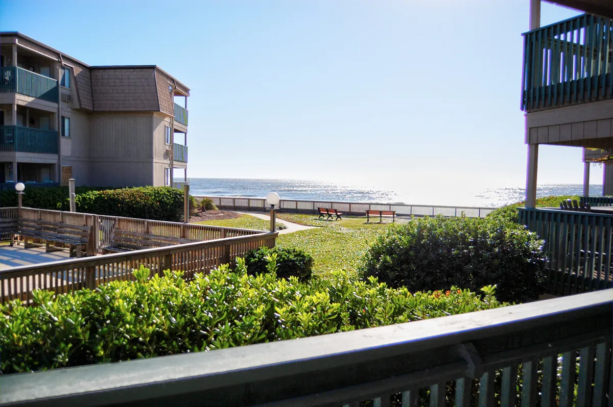 The image showcases the exterior grounds of the condo complex with lush greenery, benches inviting relaxation, and a stunning ocean view stretching out beyond, evoking a sense of tranquility. The scene suggests a place where guests can unwind, enjoy leisurely strolls by the shore, or watch the sunrise over the water.