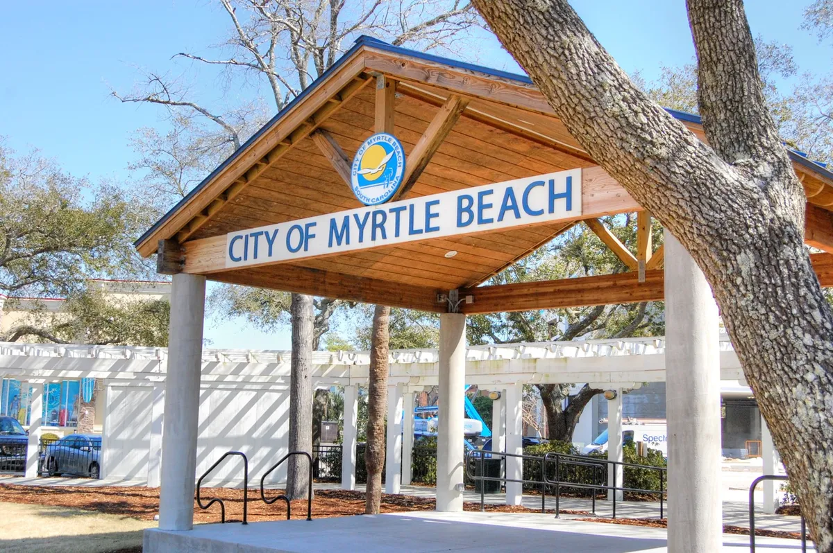 A welcoming exterior shot of the 'City of Myrtle Beach' signage set amidst a roofed structure and leafy trees. The area is designed to greet visitors, hinting at community events and social interactions. The scene is friendly and inviting, perfect for making family memories.