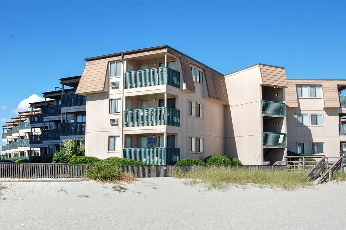 A view from the beachfront balconies looking out over a wooden railing that leads directly to the sandy beach, framed by lush green landscaping. The ocean is visible, emphasizing easy access to the shore.