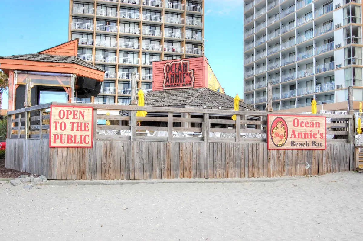 A lively beach club scene with the Ocean Annie's Beach Bar sign prominently displayed, adjacent to the beach area. There are signs welcoming the public, wooden structures, and people dining, socializing, and enjoying casual beach games at sunset.