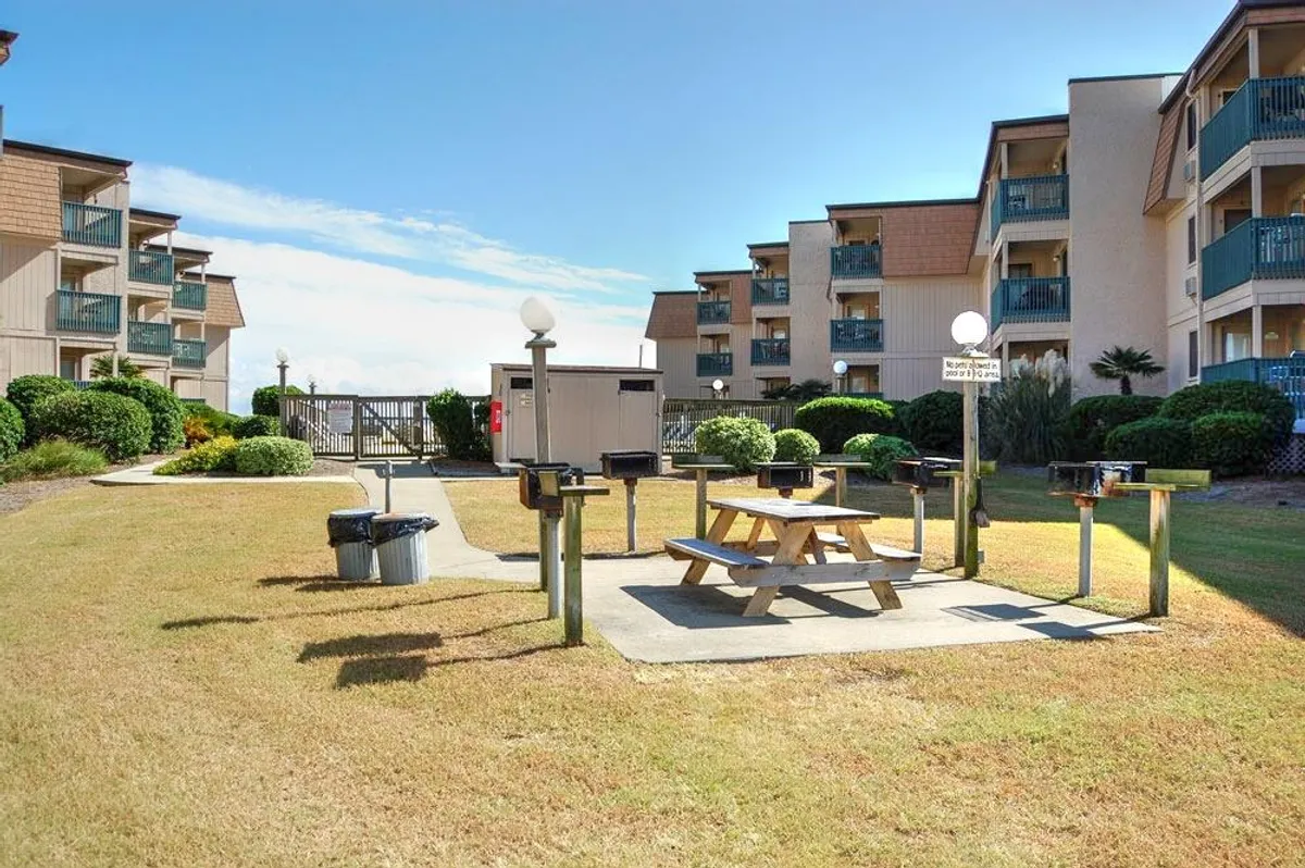 The image features a picnic table and multiple grill stations set within landscaped greenery. The area is designed for barbecuing, socializing, and enjoying meals outdoors with friends or family.
