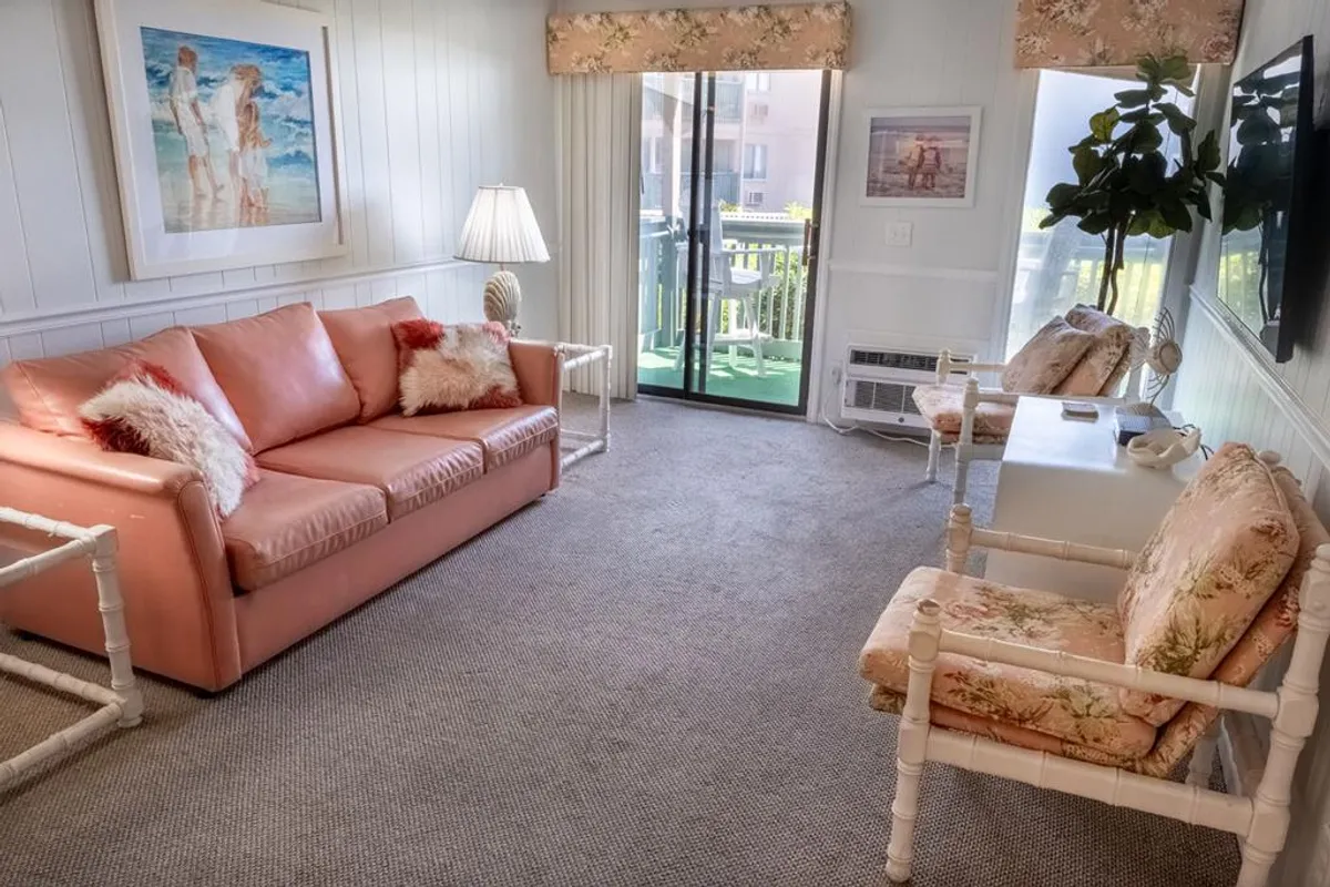 A cozy interior living space featuring a pink sofa, floral armchairs, a soft lamp glow, and a sliding glass door that opens to a private balcony. The arrangement is inviting for lounging, socializing, and enjoying intimate family gatherings. Natural light streams in, hinting at sunrise views and connection to the outside.