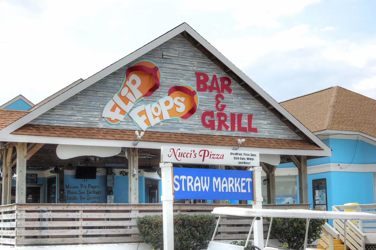 A vibrant outdoor plaza featuring the signage of Flip Flops Bar & Grill, Nucci's Pizza, and the Straw Market, suggesting options for dining, shopping, and community events. The setting feels relaxed and inviting, ideal for discovering local flavors and culture.