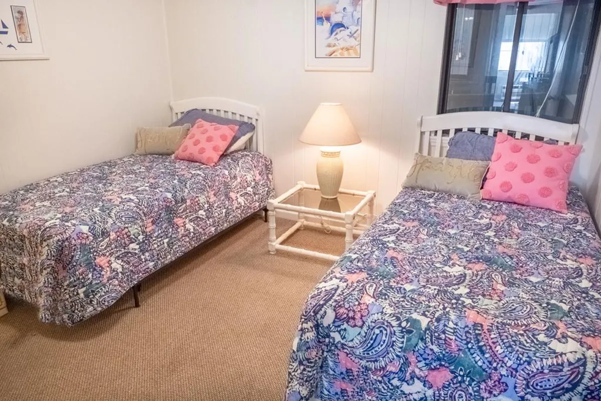 A cheerful bedroom scene featuring a comfortable bed with decorative pillows, a lamp casting warm light, and elegant wall art. The space feels inviting, bright, and ideal for families or groups, echoing the coastal theme.