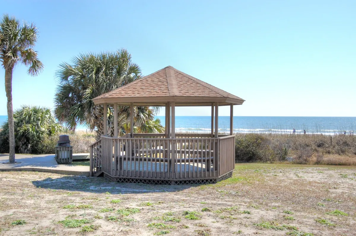 A peaceful beachfront setting featuring a charming gazebo nestled among palm trees, overlooking the expansive ocean. The scene conveys a tranquil environment, perfect for relaxation or social gatherings, with gentle morning sunlight highlighting the serene winter atmosphere.