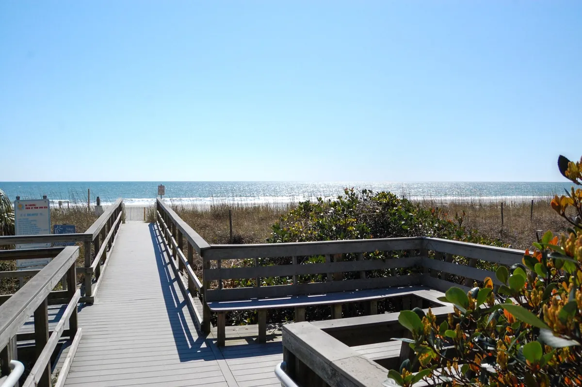 A scenic boardwalk flanked by dune grass leads directly to the sandy beach, where sunlight sparkles on the water. The area feels open, relaxed, and perfect for classic beach activities like sandcastle building and beachcombing.