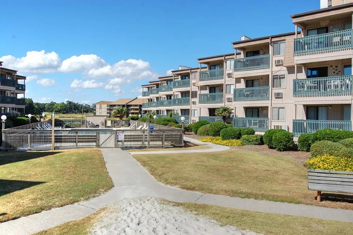 This image displays multiple balconies overlooking a walkway that leads to a pool area, surrounded by lush landscaping and under a bright blue sky. The scene feels open, welcoming, and community-oriented.
