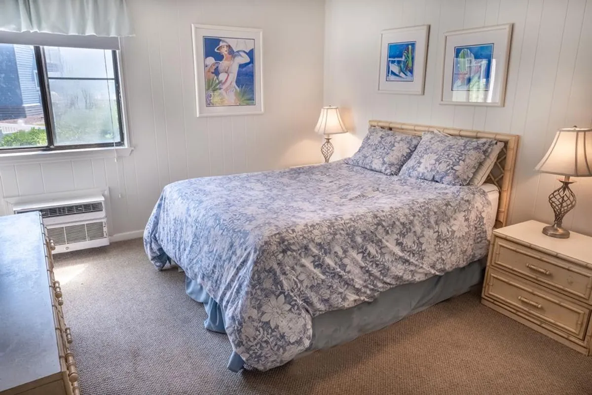 A tranquil bedroom with a plush bed adorned in floral bedding, soft lighting from decorative lamps, and calming framed artwork on the walls. The bedding and decor evoke a relaxing, coastal-inspired ambiance, ideal for unwinding after a day at the beach.